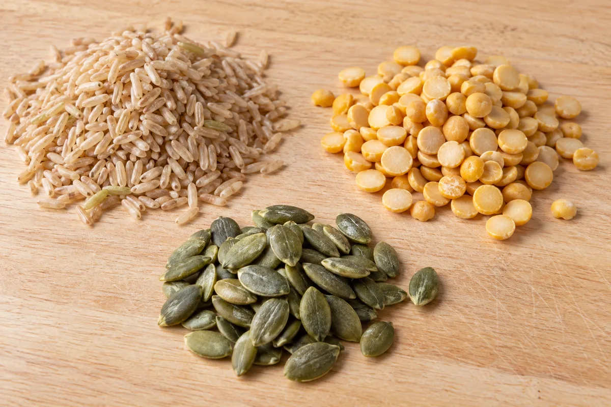 Grains de riz complet, pois cassés jaunes et graines de courge en trois petits tas sur une planche en bois clair